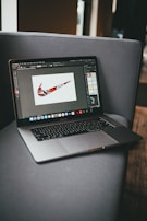 A laptop is placed on a gray couch, displaying a graphic design program with a colorful, patterned swoosh logo on the screen. The room is softly lit, with natural light coming from a window to the right.