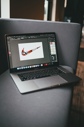 A laptop is placed on a gray couch, displaying a graphic design program with a colorful, patterned swoosh logo on the screen. The room is softly lit, with natural light coming from a window to the right.