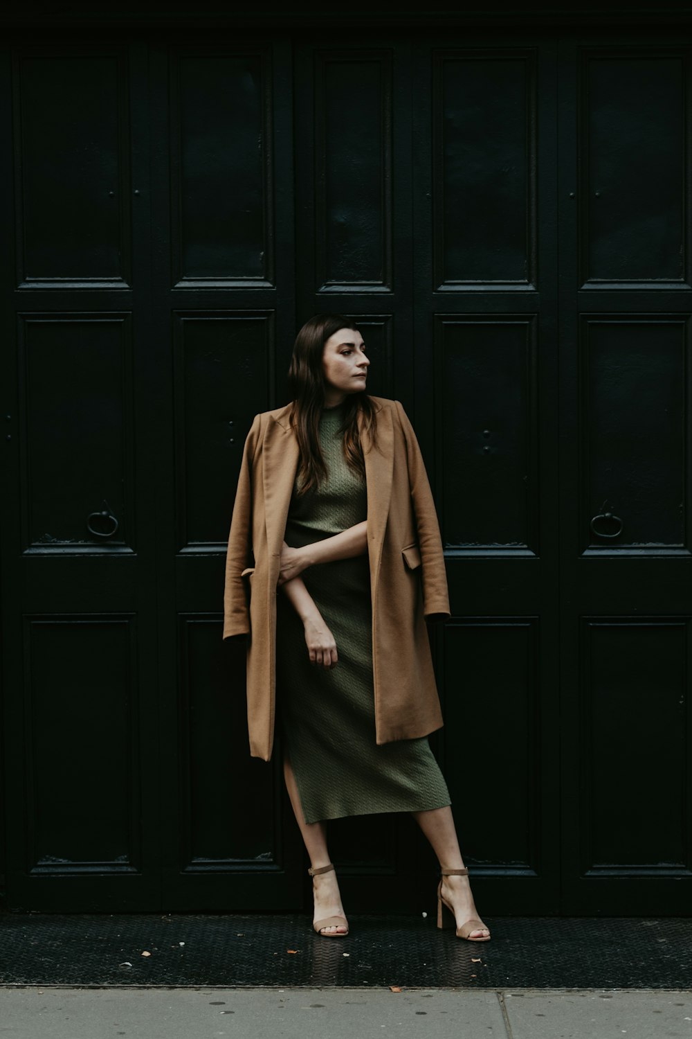 검은 나무 문 옆에 서 있는 갈색 코트를 입은 여자