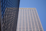 A tall skyscraper with a grid-like pattern of windows is shown from a low angle, set against a clear blue sky. The building has the logo of Wells Fargo prominently displayed on the upper portion. The building's facade reflects the neighboring structure, adding a mirrored effect.