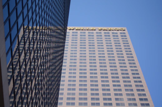 A tall skyscraper with a grid-like pattern of windows is shown from a low angle, set against a clear blue sky. The building has the logo of Wells Fargo prominently displayed on the upper portion. The building's facade reflects the neighboring structure, adding a mirrored effect.