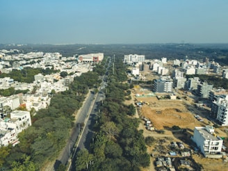 Aerial view of a cityscape with residential and commercial buildings. A long road cuts through the landscape, flanked by trees. Construction sites and cleared land indicate ongoing development. The horizon shows dense forestry beyond the urban area under a clear blue sky.