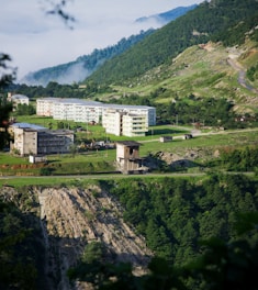 A serene landscape featuring a series of residential buildings nestled on a lush, green hillside. Mist subtly drapes over the valley in the background, adding a mysterious touch. The foreground showcases a rugged cliff with dense greenery surrounding the area.