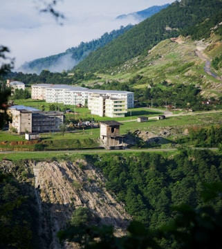 A serene landscape featuring a series of residential buildings nestled on a lush, green hillside. Mist subtly drapes over the valley in the background, adding a mysterious touch. The foreground showcases a rugged cliff with dense greenery surrounding the area.