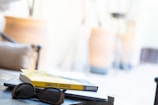 A pair of sunglasses rests on a closed laptop, with a book titled 'Cumulative Advantage' placed on top. The background is blurry, showing a light and airy environment, possibly indoors with warm tones.