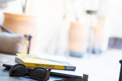 A pair of sunglasses rests on a closed laptop, with a book titled 'Cumulative Advantage' placed on top. The background is blurry, showing a light and airy environment, possibly indoors with warm tones.