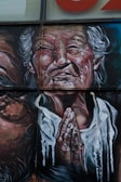 A mural depicting an elderly person with expressive, wrinkled features and gray hair. Hands are clasped together in a gesture of prayer or reflection. The intricate painting style highlights the texture of the skin and the folds of the clothing, creating an impression of wisdom and contemplation.