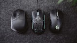 Three computer mice are placed side by side on a dark grey textured surface. The mice vary in design, with the left one having a more traditional look, the middle featuring a honeycomb pattern and LED accents, and the right one showing sleek, modern contours with a glowing logo.