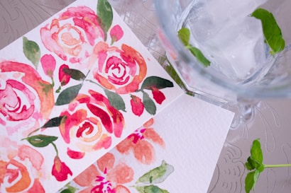 Two watercolor paintings featuring vibrant pink and red floral designs, placed on a textured surface next to a glass of water with ice and green mint leaves. The floral designs are surrounded by green leaves, creating a lively and refreshing composition.
