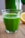 A clear glass filled with vibrant green juice sits on a wooden surface. The background includes a blurred view of a lemon and a green leafy plant.