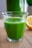 A clear glass filled with vibrant green juice sits on a wooden surface. The background includes a blurred view of a lemon and a green leafy plant.