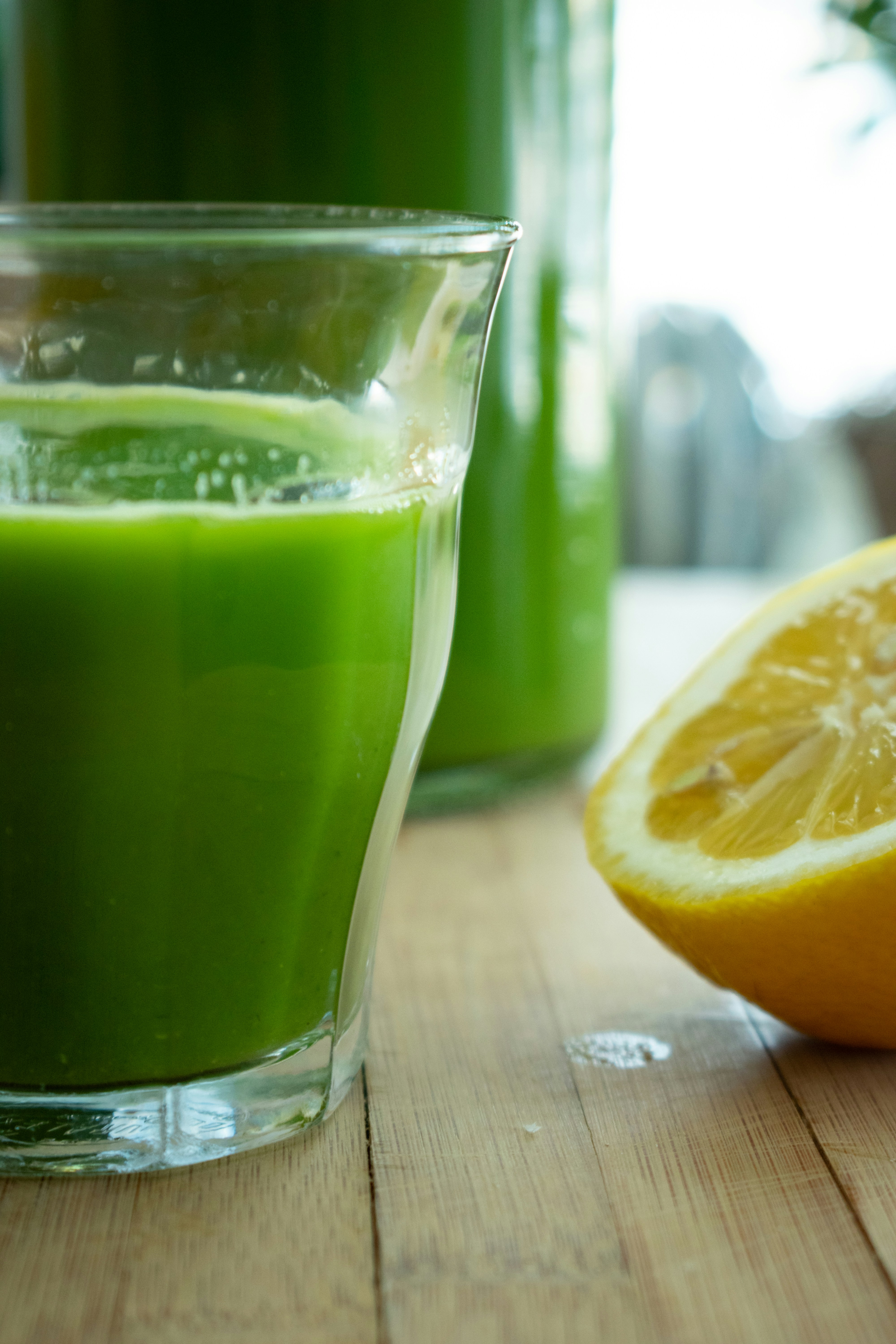 Vibrant green wheatgrass moringa spirulina shot glass surrounded by fresh green herbs and lemon on bright white surface
