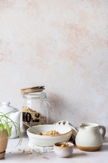 A serene kitchen scene features a bowl of cereal with a spoon, set on a light surface. Nearby, a glass jar filled with more cereal and a white teapot add to the cozy arrangement. A small potted plant adds a touch of greenery, and a cloth napkin is casually placed beside the bowl. The backdrop is a soft, neutral-toned wall.