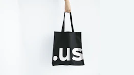 A hand holding a black tote bag with the letters '.us' printed in large, white font. The background is plain white, creating a minimalistic and clean look.