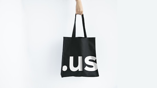 A hand holding a black tote bag with the letters '.us' printed in large, white font. The background is plain white, creating a minimalistic and clean look.
