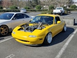 A yellow sports car with its hood removed, exposing an intricately modified engine, is parked in an outdoor parking lot. The car features wide tires and sits low to the ground, suggesting modifications for performance or aesthetic purposes. Nearby, there is another car and several other vehicles in the background.