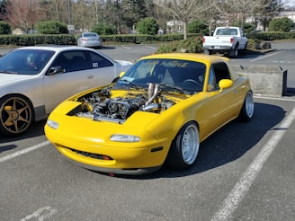 A yellow sports car with its hood removed, exposing an intricately modified engine, is parked in an outdoor parking lot. The car features wide tires and sits low to the ground, suggesting modifications for performance or aesthetic purposes. Nearby, there is another car and several other vehicles in the background.