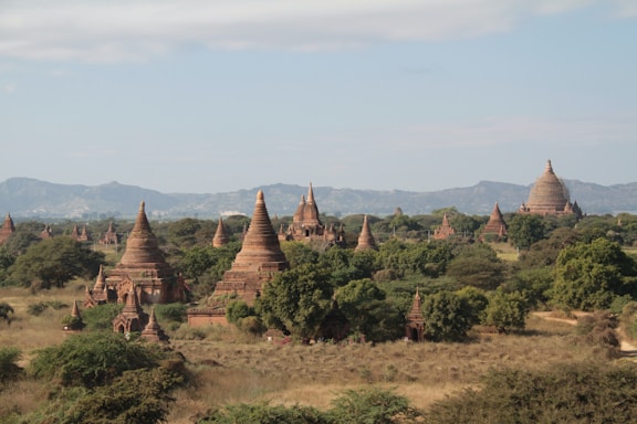 A vast, ancient landscape filled with numerous pagodas and temples, surrounded by lush greenery and hills in the distance. The structures have distinct, spire-like roofs, characteristic of traditional Southeast Asian architecture. The scene is serene and expansive with a clear sky above.