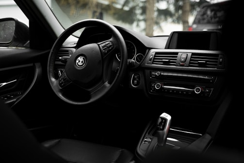 The image captures the interior of a car highlighting the steering wheel and dashboard. The steering wheel features the logo of a well-known automobile brand. The dashboard includes air vents, control buttons, and an infotainment screen, all designed with sleek, modern aesthetics. The interior appears to be made of high-quality, dark-colored materials.