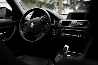 The image captures the interior of a car highlighting the steering wheel and dashboard. The steering wheel features the logo of a well-known automobile brand. The dashboard includes air vents, control buttons, and an infotainment screen, all designed with sleek, modern aesthetics. The interior appears to be made of high-quality, dark-colored materials.
