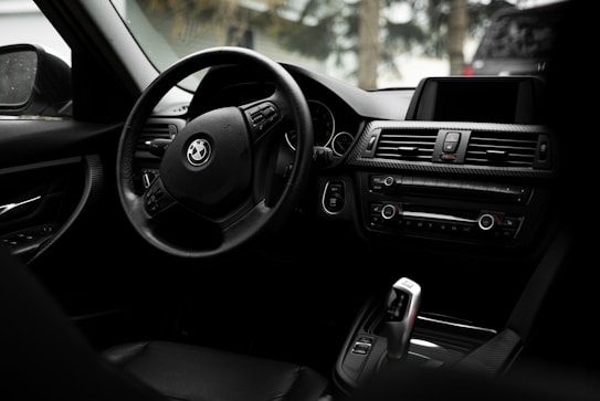 The image captures the interior of a car highlighting the steering wheel and dashboard. The steering wheel features the logo of a well-known automobile brand. The dashboard includes air vents, control buttons, and an infotainment screen, all designed with sleek, modern aesthetics. The interior appears to be made of high-quality, dark-colored materials.