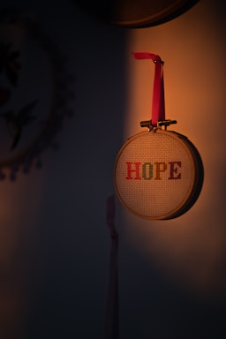 A wooden embroidery hoop hangs on a wall, containing cross-stitching that spells out the word 'HOPE' in colorful letters. A red ribbon is attached to the hoop. Soft warm light casts a shadow, creating a cozy and intimate atmosphere.
