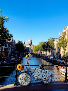 A decorated bicycle covered with a daisy-patterned cloth and some sunflowers is parked on a bridge overlooking a canal. The scene is set in a picturesque cityscape featuring rows of traditional European houses along the waterway. Boats are docked alongside the canal, while trees line the street. The sky above is clear and bright blue.