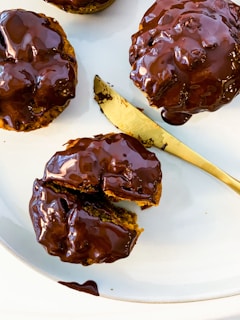 Three chocolate-glazed pastries are arranged on a white plate with a gold knife. The pastries are round and richly covered in glistening chocolate, with one of them partially sliced open to reveal a soft interior.