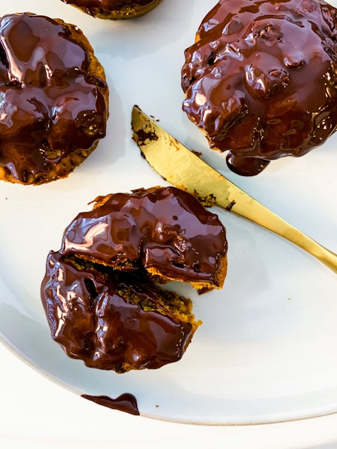 Three chocolate-glazed pastries are arranged on a white plate with a gold knife. The pastries are round and richly covered in glistening chocolate, with one of them partially sliced open to reveal a soft interior.