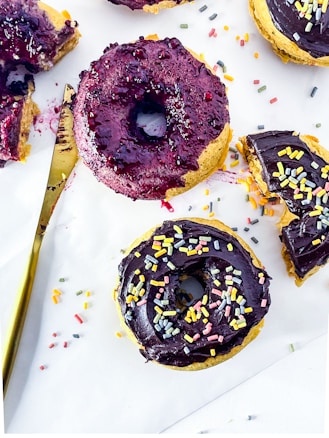 Two doughnuts with colorful sprinkles are placed on a white surface. One doughnut is coated in glossy chocolate frosting and the other in a rich, berry glaze. A gold knife with traces of berry glaze lays nearby, and loose sprinkles are scattered around.