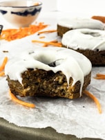 A close-up of a frosted donut with a bite taken out of it, resting on a piece of parchment paper. Shredded carrots are scattered around, and in the background, there is a bowl with a blue pattern.