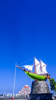 A large sculpture of a traditional sailing ship is mounted on a stone pedestal against a clear blue sky. The ship is vibrant green with white sails and red details. The background features a mosque with red and white domes, and large white letters spell out 'LOSARI' on the left.