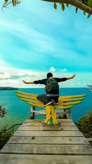 A person with a backpack sits with arms outstretched on a wooden platform shaped like wings, overlooking a vibrant turquoise sea and distant land. The sky is clear and bright, adding to the serene ambiance.