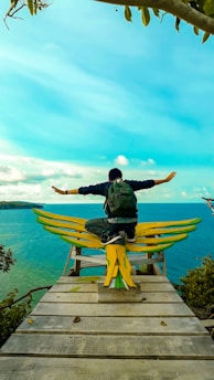 A person with a backpack sits with arms outstretched on a wooden platform shaped like wings, overlooking a vibrant turquoise sea and distant land. The sky is clear and bright, adding to the serene ambiance.