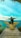 A person with a backpack sits with arms outstretched on a wooden platform shaped like wings, overlooking a vibrant turquoise sea and distant land. The sky is clear and bright, adding to the serene ambiance.