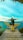 A person with a backpack sits with arms outstretched on a wooden platform shaped like wings, overlooking a vibrant turquoise sea and distant land. The sky is clear and bright, adding to the serene ambiance.