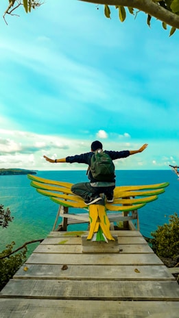 A person with a backpack sits with arms outstretched on a wooden platform shaped like wings, overlooking a vibrant turquoise sea and distant land. The sky is clear and bright, adding to the serene ambiance.