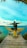 A person with a backpack sits with arms outstretched on a wooden platform shaped like wings, overlooking a vibrant turquoise sea and distant land. The sky is clear and bright, adding to the serene ambiance.