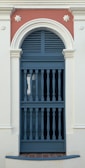 An ornate door features a semi-circular archway with decorative molding. The door itself has a series of vertical bars, typical of colonial architectural style, and is painted in a muted blue shade. The surrounding wall is off-white, with a contrasting terracotta-colored section above the door, accentuated by floral motifs.