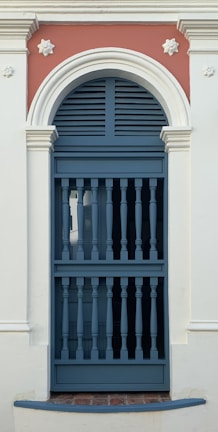 An ornate door features a semi-circular archway with decorative molding. The door itself has a series of vertical bars, typical of colonial architectural style, and is painted in a muted blue shade. The surrounding wall is off-white, with a contrasting terracotta-colored section above the door, accentuated by floral motifs.