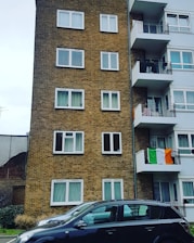 A multi-story residential building is seen with a brick facade on the left and white balconies on the right. Multiple windows are visible on both sides of the building. An Irish flag is draped over one of the balconies. In the foreground, two parked cars are visible. The surrounding environment includes a partially visible fence and some greenery.