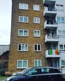 A multi-story residential building is seen with a brick facade on the left and white balconies on the right. Multiple windows are visible on both sides of the building. An Irish flag is draped over one of the balconies. In the foreground, two parked cars are visible. The surrounding environment includes a partially visible fence and some greenery.