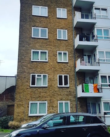 A multi-story residential building is seen with a brick facade on the left and white balconies on the right. Multiple windows are visible on both sides of the building. An Irish flag is draped over one of the balconies. In the foreground, two parked cars are visible. The surrounding environment includes a partially visible fence and some greenery.