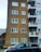 A multi-story residential building is seen with a brick facade on the left and white balconies on the right. Multiple windows are visible on both sides of the building. An Irish flag is draped over one of the balconies. In the foreground, two parked cars are visible. The surrounding environment includes a partially visible fence and some greenery.