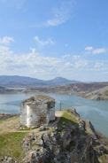 A small, rustic stone chapel stands atop a rocky cliff overlooking a serene lake. Surrounding the chapel are rolling hills and distant mountains under a mostly clear sky with some scattered clouds. A cross is positioned near the chapel, enhancing the peaceful and contemplative atmosphere of the landscape.