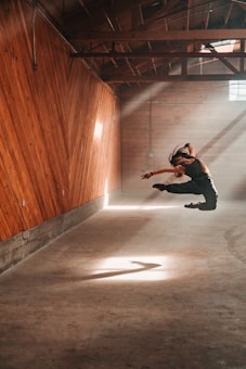 A dancer captured mid-air in a rustic wooden room with slanted walls. Sunlight streams through a window, casting dramatic shadows on the floor and walls. The scene is filled with dynamic motion and a sense of freedom.