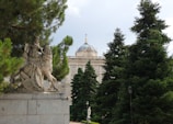 A statue of a medieval knight on horseback is prominently situated on a stone pedestal amidst lush green pine trees. In the background, there is an ornate building with a dome topped with a cross, exhibiting classical architecture.