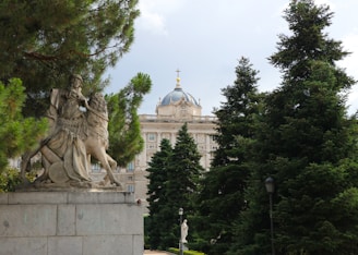 A statue of a medieval knight on horseback is prominently situated on a stone pedestal amidst lush green pine trees. In the background, there is an ornate building with a dome topped with a cross, exhibiting classical architecture.