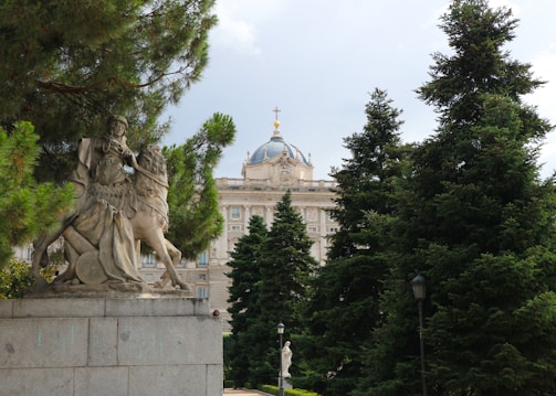 A statue of a medieval knight on horseback is prominently situated on a stone pedestal amidst lush green pine trees. In the background, there is an ornate building with a dome topped with a cross, exhibiting classical architecture.