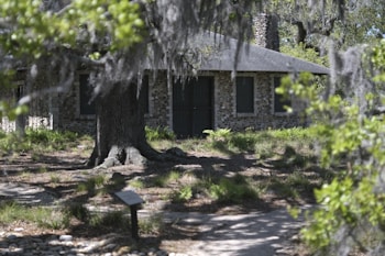 A rustic stone cottage is surrounded by lush greenery and Spanish moss-draped trees, creating a tranquil and secluded atmosphere. The cottage has a small chimney and is partly obscured by the large tree in the foreground. Sunlight filters through the leaves, casting dappled shadows on the ground.